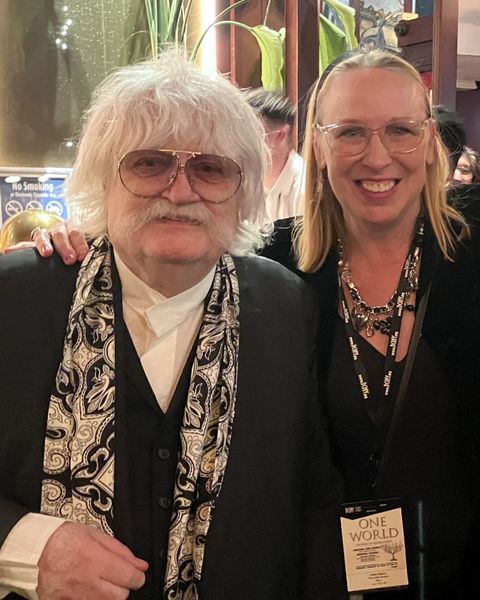 Lorilee Bajema smiling beside Sir Karl Jenkins, who wears tinted glasses and a patterned scarf, at the One World concert
