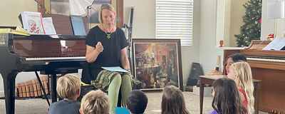 Lorilee Bajema seated at her grand piano, talking with six children sitting on the carpet in front of her