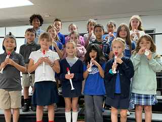 A class of children in school uniforms, each holding a recorder, smiling on the risers