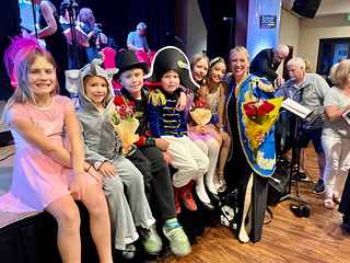 Children in costume sitting along the edge of a stage beside Lorilee, who wears a blue military-style jacket and holds a bouquet