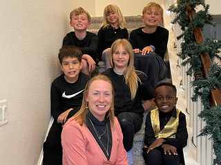 Lorilee sitting on a staircase with six smiling children dressed in black, one in a gold sequin vest