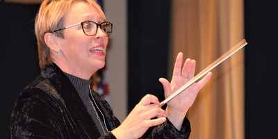 Lorilee Bajema conducting with a baton, mid-phrase and smiling