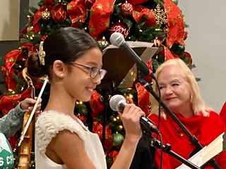 A young girl in a white dress singing into a microphone beside a decorated Christmas tree